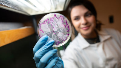 young woman in lab coat blurred behind her gloved hand holding a petri dish